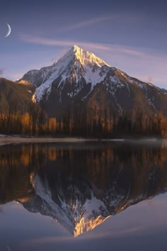 a mountain is reflected in the still waters of a lake at sunset with a half moon