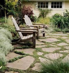 two wooden chairs sitting on top of a stone walkway next to a house and bushes