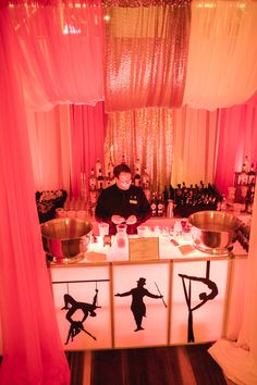 a man standing in front of a counter with various items on it and pink curtains