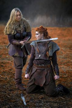 two women dressed in medieval costumes holding swords and looking at each other while sitting on the ground