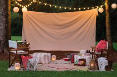 an outdoor picnic with lights strung from the trees and blankets on the ground, lanterns in the background