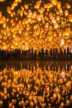 many lit up lanterns floating in the air over a body of water with people standing around