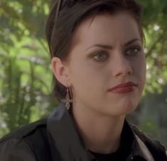 a woman with dark hair and green eyes looks into the distance while standing in front of trees