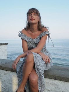 a woman sitting on the edge of a wall next to the ocean with her legs crossed