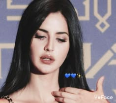 a woman with long black hair holding her hand up to her chest and wearing a blue heart shaped brooch