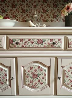 a white dresser with flowers painted on the top and bottom, in front of a floral wallpaper