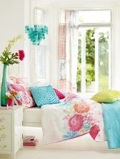 a bed with colorful pillows and blankets in a white room next to a large window