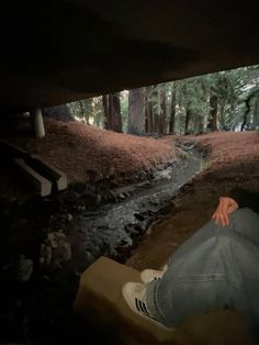 a person sitting on the ground with their feet up in front of a creek and trees