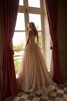 a woman standing in front of a window wearing a wedding dress and looking out the window