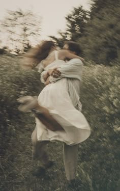 two women in white dresses are hugging each other while walking through the grass with trees and bushes behind them