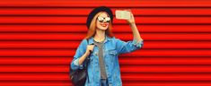 Portrait of happy smiling young woman taking selfie with smartphone wearing jean jacket, black round hat on red background stock photo Round Hat, Red Background, Jean Jacket, Hats, Red, How To Wear, Black