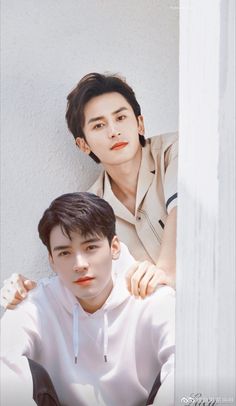 two young men sitting next to each other in front of a white wall, one with his arm around the other's shoulder