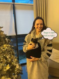 a woman holding a cat in front of a christmas tree with a thought bubble above her head