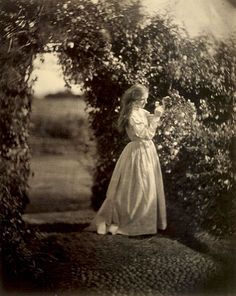 an old black and white photo of a woman in a dress standing under a tree