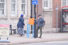 two people standing on the sidewalk next to a pole with a bike rack attached to it