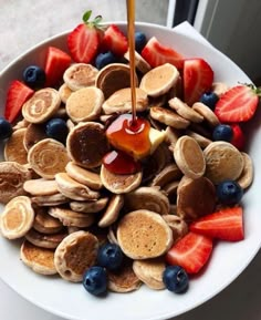 a white plate topped with pancakes and fruit next to a cup of syrup being poured on top