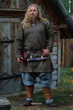 a man with long hair and beard standing in front of a wooden building wearing an old fashioned outfit