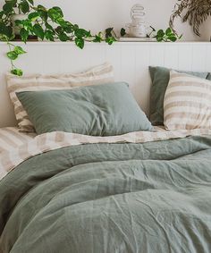 an unmade bed with green sheets and pillows in a white room next to potted plants