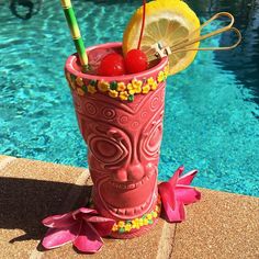 a pink tiki style drink next to a swimming pool with flowers and fruit in it