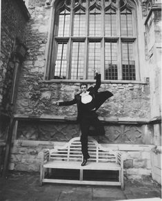 black and white photograph of man jumping on bench in front of building with large window