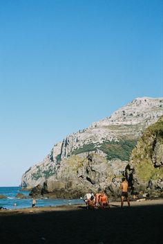 some people are standing on the beach with mountains in the background