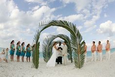 a bride and groom standing under a palm tree on the beach with their wedding party