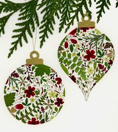 two ornaments hanging from a tree branch with green leaves and red flowers on it's ornament