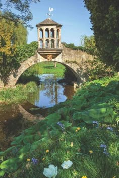 an old stone bridge over a small stream in the middle of a park with flowers and trees