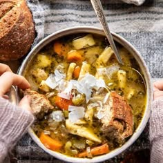 two hands holding a bowl of soup with bread on the side