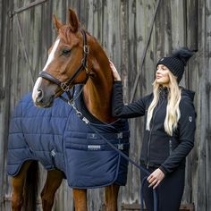 a woman standing next to a brown horse wearing a blue blanket on it's back