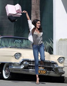 a woman is jumping in the air with a box on her hand and an old car behind her