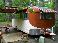 an orange and white camper parked under a canopy in the woods next to picnic tables