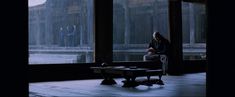 a person sitting on a bench in front of large windows with rain coming through them