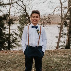 a young man wearing suspenders and a bow tie standing in front of a lake