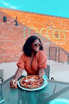 a woman sitting at an outdoor table with a pizza in front of her, smiling