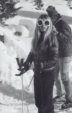 two people standing in the snow with skis on and one person wearing goggles