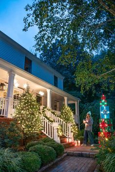 a person standing on a porch next to christmas trees and lights in front of a house