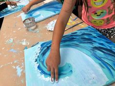 two children are painting on canvass with blue and white paint, while one child is holding their hand out to the other