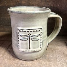 a ceramic mug with a dragonfly design on the front and sides, sitting on a wooden surface