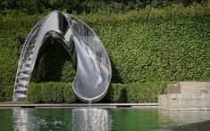 a water slide that is in the middle of a pool with bushes behind it and grass on either side