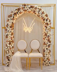 two white chairs sitting next to each other under a floral arch with lights on it