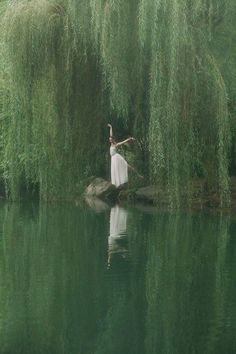 a woman in a white dress is standing on a rock by the water with willow branches