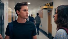 a young man and woman standing in a hallway next to lockers looking at each other