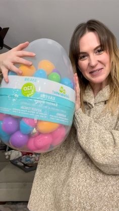 a woman holding up a large plastic container filled with colorful balls in her hands and smiling at the camera