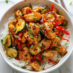 a white bowl filled with chicken and veggies on top of rice next to chopsticks