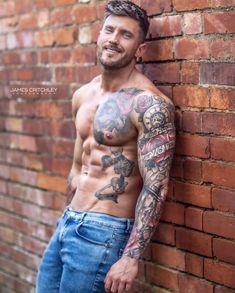 a shirtless man leaning against a brick wall with tattoos on his arms and chest