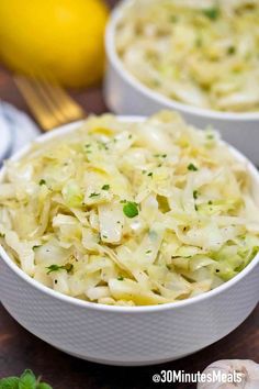 two white bowls filled with coleslaw on top of a wooden table next to lemons