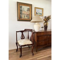 a chair sitting next to a lamp on top of a wooden dresser under a painting