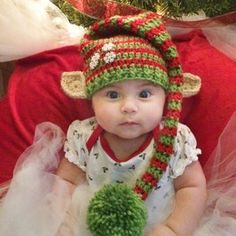 a baby wearing a knitted elf hat sitting in front of a christmas tree