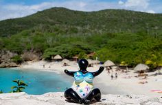 a statue sitting on top of a sandy beach next to the ocean in front of a lush green hillside